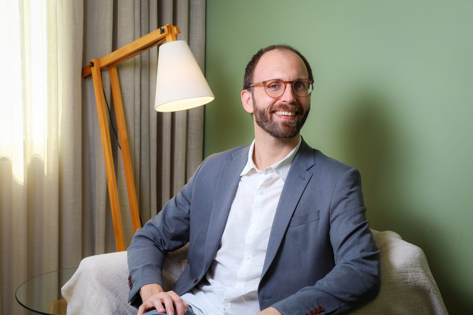 Dr. Roberto Gimena sorrindo na poltrona do consultório, com a luminária e a cortina ao lado