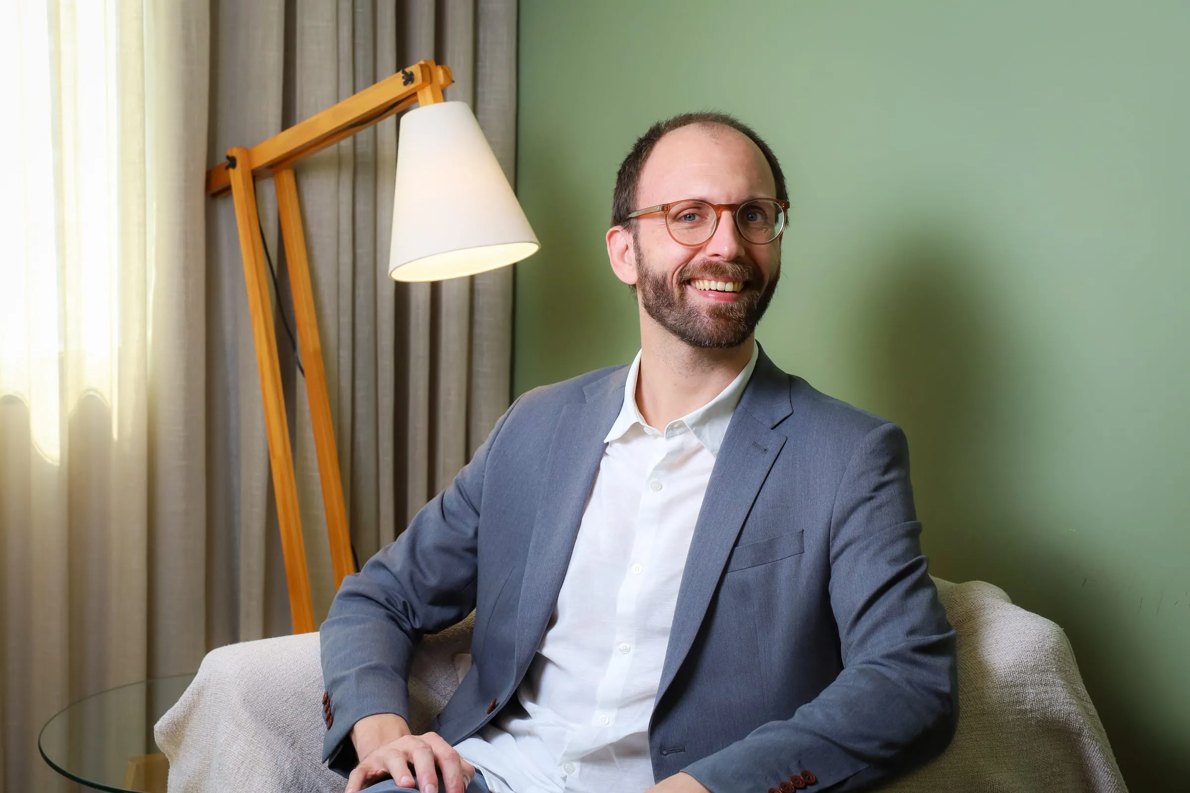 Dr. Roberto Gimena sorrindo na poltrona do consultório, com a luminária e a cortina ao lado